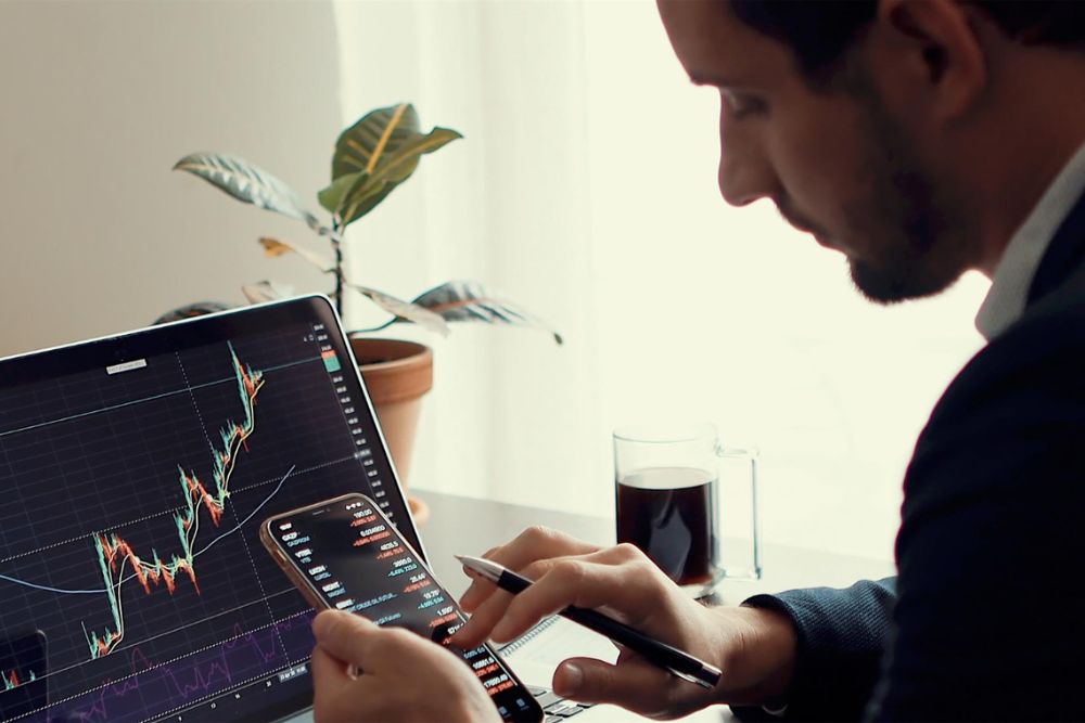 investor sedící s mobilem u PC a sledující vývoj akcií